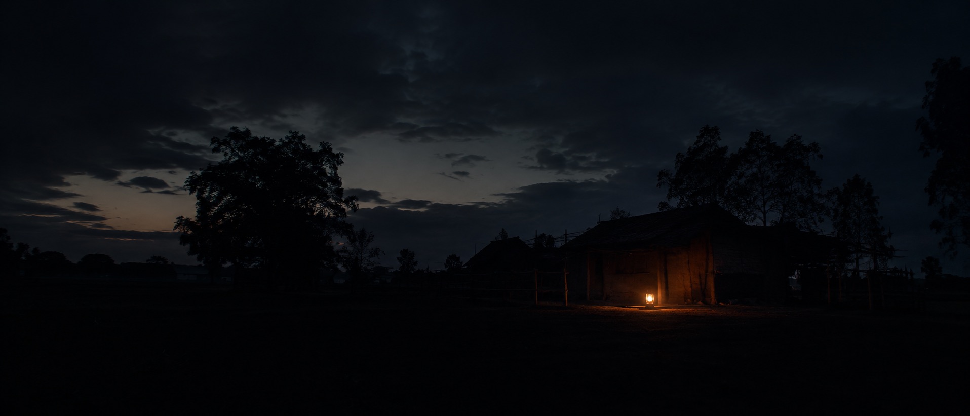 A village silhouette at deep dusk with a single kerosene lantern as the only light